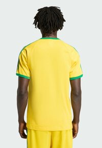 Osoba o ciemnej karnacji i dredach ubrana w żółty strój sportowy z zielonym obszyciem, stoi tyłem na tle jednolitego jasnego tła.