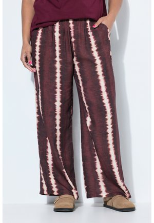 Kvinde iført vinrøde og beige brede bukser med striber og lommer samt beige slip-on sko, stående op ad en hvid væg.