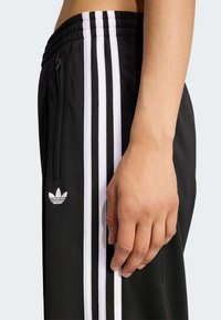 Czarne spodnie sportowe z trzema białymi pionowymi paskami, kieszenią na zamek, prążkowanym paskiem w talii oraz haftowanym logo po lewej stronie.