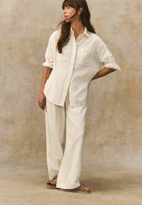 Souprava lněné košile a kalhot v off-white s jemnými vertikálními pruhy. Obsahuje košili s knoflíkovým zapínáním a dvěma náprsními kapsami a kalhoty s uvolněným střihem.