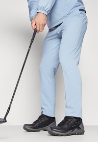 Világoskék golfnadrág, texturált felülettel és tapered fazonnal, fekete golfcipővel, amely robusztus talppal és zsinóros részletekkel rendelkezik.