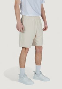 Pantaloni sportivi beige con elastico in vita, dotati di tasche laterali e texture morbida, abbinati a sneaker bianche e calzini chiari.