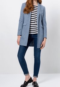 Vaaleansininen bleiseri, jossa on kaksi etutaskua, yhdistetty musta-valkoraitapaitaan, sinisiin farkkuihin ja mustiin loafereihin.