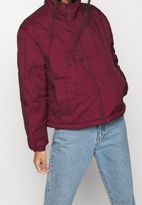 Burgundy vadderad jacka med hög krage, prydd med ett blommönster, två sidofickor och dragkedja. Bärs över ljusblå jeans.