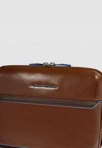 Bruine leren tas met een voorvak met ritssluiting, blauwe accentstiksels en een zilveren logo dat in het oppervlak is geperst. Gladde textuur.