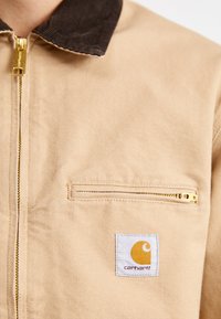 Jachetă de lucru bej cu fermoar în față, guler din catifea maro și un buzunar pe piept. Prezintă un eticheta Carhartt cu un logo galben. Material texturat.