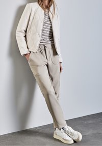 Ljust beige quiltad bomberjacka över en randig skjorta, med beiga cargobyxor med fickor och vita sneakers med detaljer.