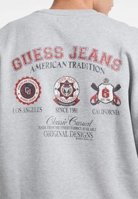 Felpa grigia con caratteri grafici audaci in rosso e nero, loghi e date di fondazione. Il design mette in risalto "Guess Jeans" e la tradizione americana.