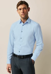 Chemise bleu clair à manches longues avec un motif texturé, devant boutonné et boutons sombres contrastés, assortie à un pantalon sombre et une ceinture.
