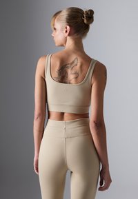 Kvinne med blondt hår i en samlede, iført beige sports-bh og leggings, som viser en stor drage-tatovering på øvre del av ryggen mot en grå bakgrunn.