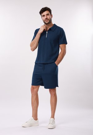 Άνδρας που φοράει μπλε σκούρο πουκάμισο με κοντά μανίκια και φερμουάρ και ταιριαστά σορτς με λευκά αθλητικά, μπροστά σε απλό φόντο.