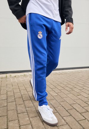 Person, die blaue Real Madrid Trainingshose mit weißen Streifen und weiße Turnschuhe auf einer gepflasterten Fläche trägt.