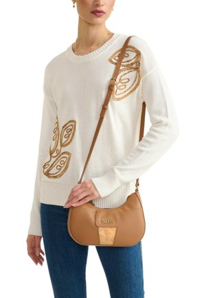 Donna con maglione bianco ricamato con foglia marrone, che tiene una piccola borsa a tracolla in pelle marrone chiaro con logo del marchio visibile.