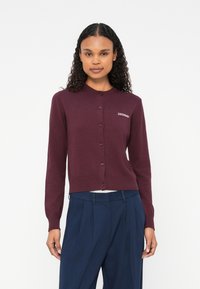 Cardigan color marrone in morbido filato con scollo tondo, bottoni frontali e orlo a coste. Presenta un logo ricamato sul lato sinistro.