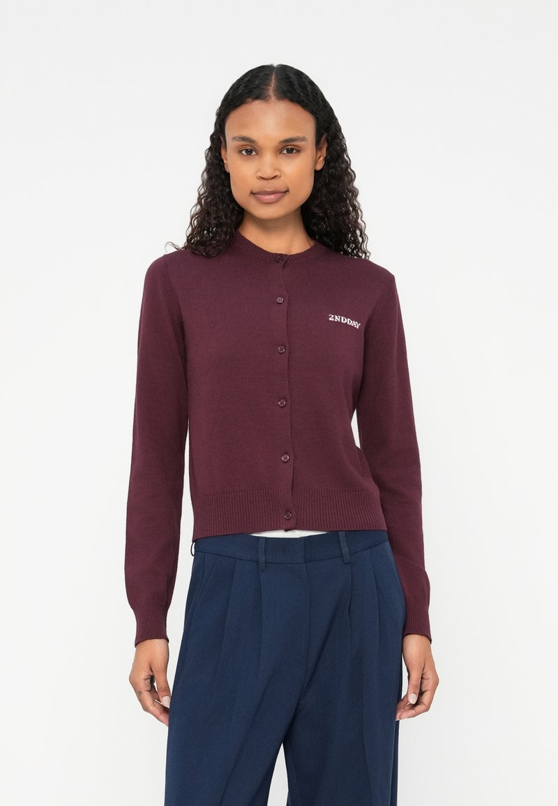 Cardigan color marrone in morbido filato con scollo tondo, bottoni frontali e orlo a coste. Presenta un logo ricamato sul lato sinistro.