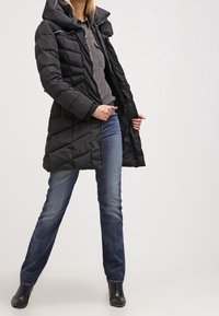 Abrigo de plumas negro con capucha, cierre frontal y diseño acolchado. Combinado con una camiseta gris y jeans azules, mostrando un look casual.