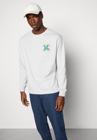 Camisa cinza de manga longa com um logo verde e um design de louros no peito esquerdo, combinada com jeans de denim azul e um boné branco.