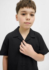 Camisa negra de manga corta con un patrón texturizado, cuello tipo polo y cierre frontal de botones, sostenida por la mano de un niño.