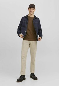Veste en denim avec des boutons, pull en tricot marron, pantalon beige à jambes droites et bottes noires à lacets. Fond gris.