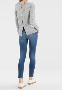Un pull gris clair avec une découpe en forme de clé et trois nœuds à l'arrière, associé à un jean skinny bleu et des baskets blanches.