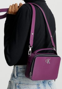 Osoba w jasnoniebieskich dżinsach i czarnej bluzce, niosąca fioletową torebkę typu crossbody marki Calvin Klein z srebrnymi akcentami i regulowanym paskiem.