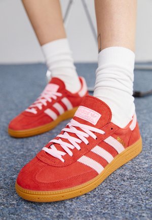 Punaiset Adidas-tossut, joissa valkoiset raidat ja nauhat, valkoisten sukkien kanssa sinisellä matolla, jossa näkyy tatuoitu nilkka.