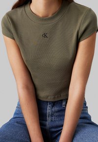 Olivově zelený žebrovaný crop top s kulatým výstřihem a krátkými rukávy. Na hrudi je černé logo "CK". Detail texturované tkaniny.