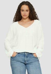 Witte, losvallende blouse met een V-hals, lange mouwen en een subtiele textuur. Gecombineerd met blauwe jeans.