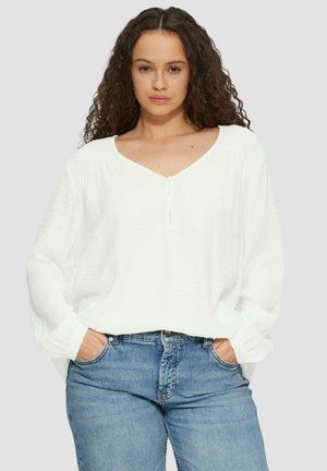 Witte, losvallende blouse met een V-hals, lange mouwen en een subtiele textuur. Gecombineerd met blauwe jeans.