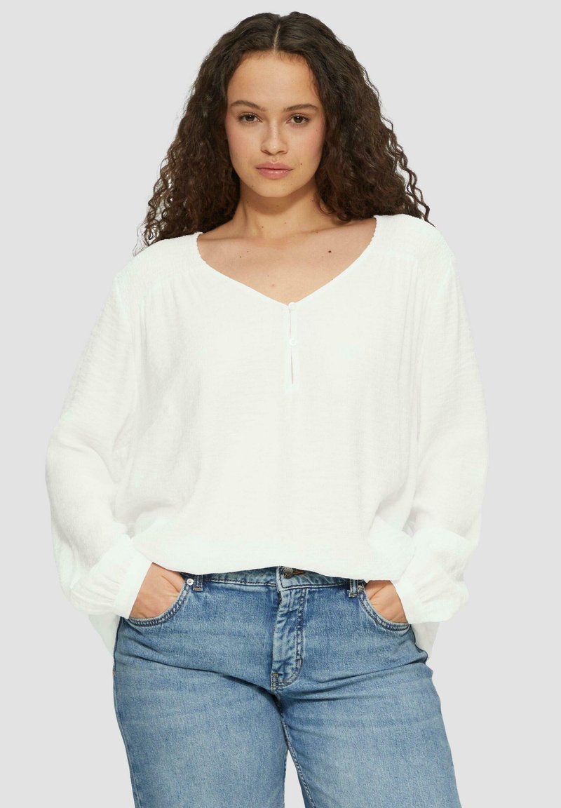 Witte, losvallende blouse met een V-hals, lange mouwen en een subtiele textuur. Gecombineerd met blauwe jeans.