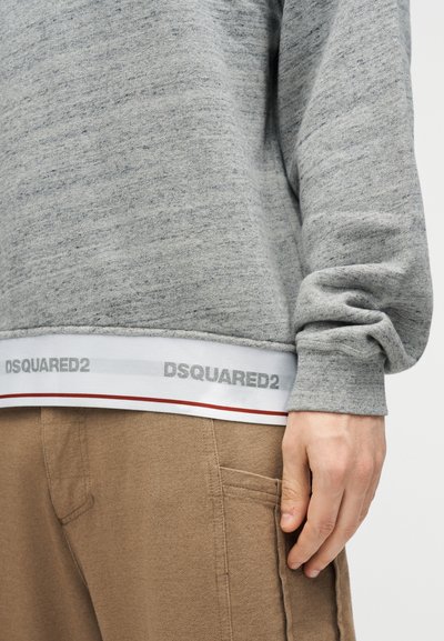 Persona che indossa un maglione grigio con il logo DSQUARED2 sull'orlo e pantaloni marrone chiaro con tasca visibile e mano posata al lato.
