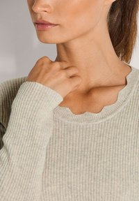 Die Hand einer Frau berührt ihren Hals, während sie einen hellbeigen, gerippten Pullover mit gewelltem Halsausschnitt trägt.