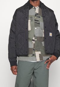Zwarte gewatteerde jas met geribbelde manchetten, gecombineerd met een overhemd met meerdere patronen in groene tinten, met knoopsluiting en borstzakken.