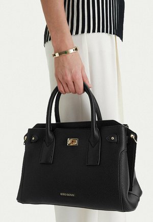 Mano che tiene una borsa a tracolla in pelle nera testurizzata con chiusura e logo dorati, indossando un braccialetto dorato e pantaloni bianchi con top a strisce nere.