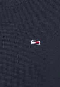 Marineblauer Strickpullover mit geripptem Ausschnitt und einem kleinen, auf der Brust gestickten Logo mit roten, weißen und blauen Akzenten.