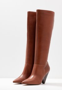 Bottes hautes en cuir marron jusqu'aux genoux avec un bout pointu et un talon conique. Texture lisse avec un design épuré et des détails de couture minimalistes.