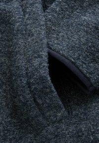 Marineblauw, gestructureerde wollen stof met een pluizige oppervlakte; bevat een zijzak gevoerd met donkere materialen, wat het algehele ontwerp versterkt.