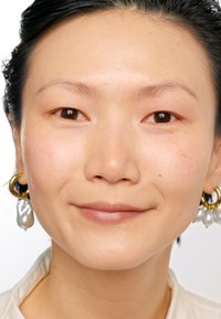 Gros plan sur un visage aux traits lisses et à la peau claire. Présence de boucles d'oreilles créoles avec des accents noirs et de grands détails en forme de perles. Couleur de lèvres neutre.