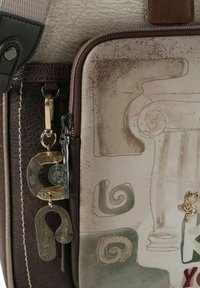 Strukturierte Lederhandtasche mit beige-braunem Farbschema. Verziert mit dekorativen Wirbeln, einem Katzenanhänger und goldfarbenen Beschlägen.