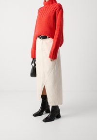 Roter gestrickter Rollkragenpullover, cremefarbener Midirock mit diagonaler Naht, schwarzer Gürtel, schwarze Stiefeletten mit quadratischer Spitze und Absatz, kleine schwarze Handtasche.