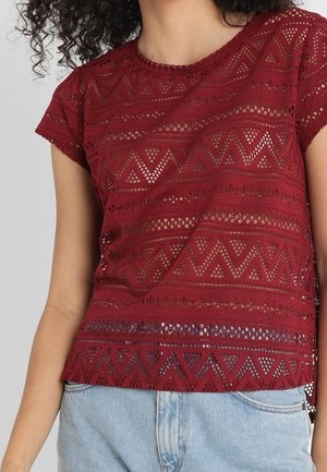 Kobieta w bordowej koronkowej bluzce z krótkim rękawem i geometrycznymi wzorami oraz jasnoniebieskich dżinsach, widoczna od klatki piersiowej do bioder.