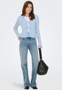 Cardigan bleu clair en maille avec un détail noué à l'avant, associé à un jean évasé bleu clair et un sac à main noir.