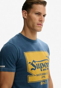 Blauw katoenen T-shirt met een grote gele afbeelding en tekst "Superdry Motorbikes JPN." Ronde hals, korte mouwen, casual pasvorm.