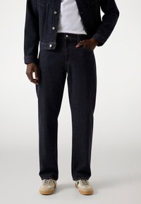 Dark denim jeans with a straight-leg cut, featuring orange stitching, five pockets, and a contrasting waistband. Worn with beige sneakers.