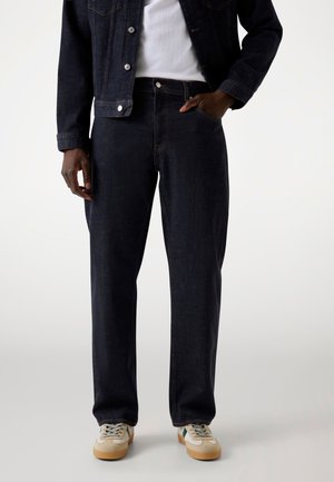 Dark denim jeans with a straight-leg cut, featuring orange stitching, five pockets, and a contrasting waistband. Worn with beige sneakers.