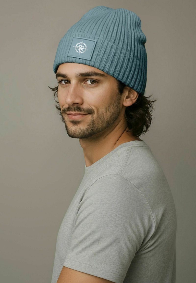 Blue ribbed knit beanie with a stitched compass patch, worn by a person in a light gray shirt, facing slightly left. Soft texture.