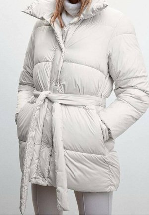 Femme portant une doudoune claire ceinturée à col montant, par-dessus un pull à col roulé blanc, les mains dans les poches.