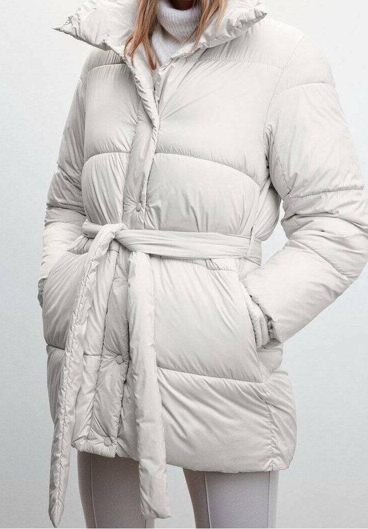 Femme portant une doudoune claire ceinturée à col montant, par-dessus un pull à col roulé blanc, les mains dans les poches.