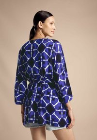 Tunik a motivi blu e bianchi con cintura, caratterizzata da maniche lunghe e una vestibilità rilassata, abbinata a pantaloncini in denim. Texture del tessuto liscia.