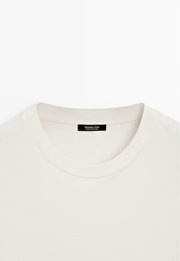 T-shirt di cotone color crema con collo rotondo, caratterizzato da un colletto a coste e un'etichetta nera cucita all'interno del collo con del testo.